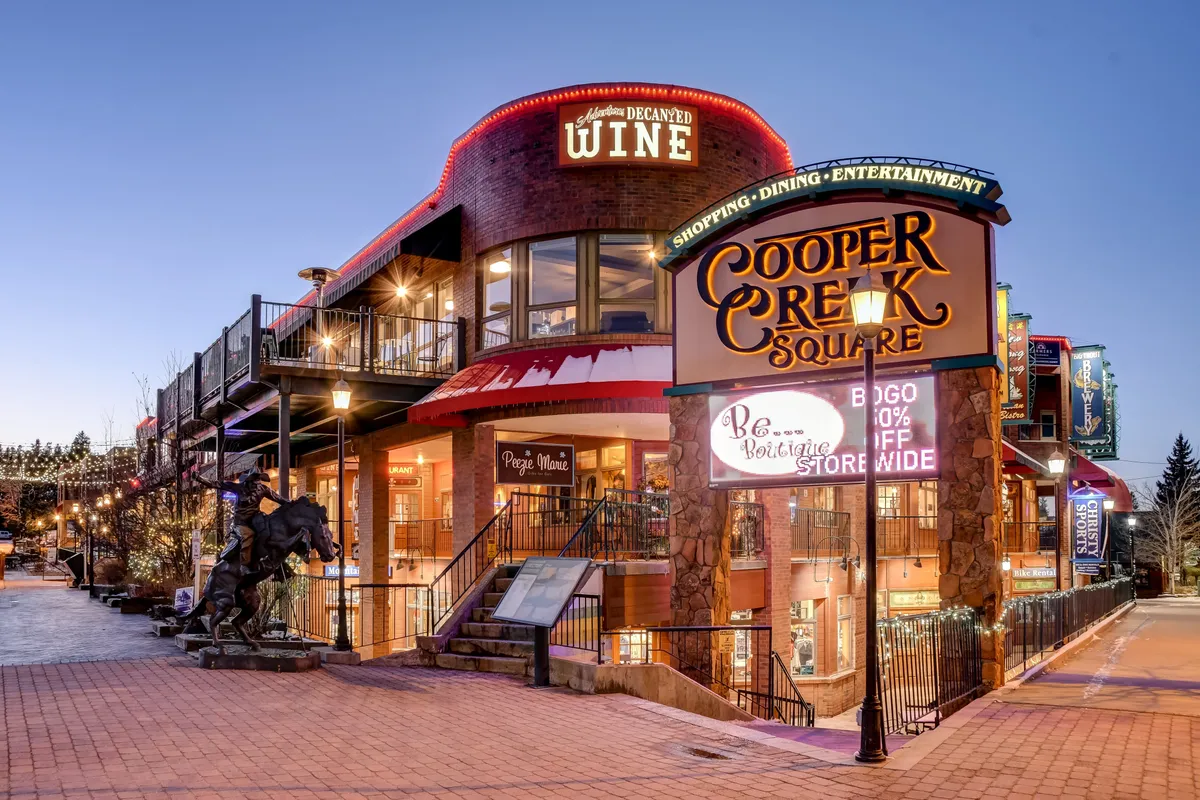 A bright, bustling scene at Cooper Creek Square in downtown Winter Park, highlighting the recognizable sign, outdoor seating, and families enjoying the area. The lively atmosphere includes shops and a horse statue, capturing the sense of community and entertainment.