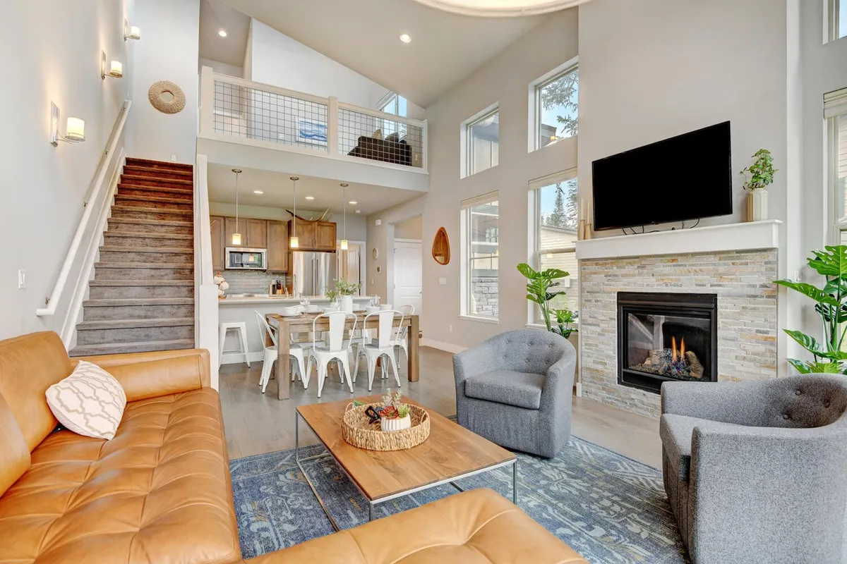 The image showcases a stylish, open-concept living area featuring a plush leather sofa facing a warm stone fireplace, with a modern dining area just beyond. Expansive windows frame mountain views and fill the space with natural light, while a staircase hints at more space to explore. The setting is perfect for cozy family gatherings or relaxing after a day of adventure.