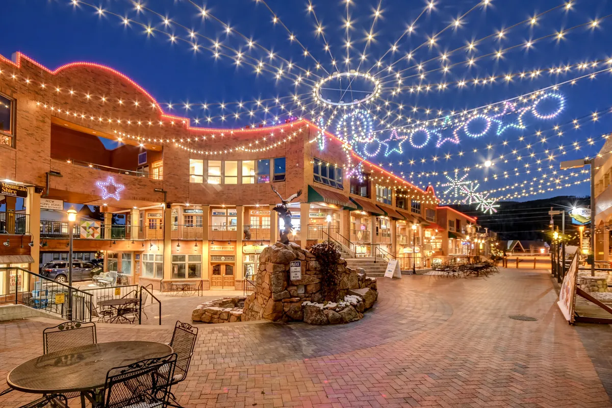 An inviting outdoor shopping and dining area, illuminated by decorative string lights, with a stone fountain and bustling activity in the background. The setting exudes a festive, social vibe ideal for evening gatherings and experiences in downtown Winter Park.