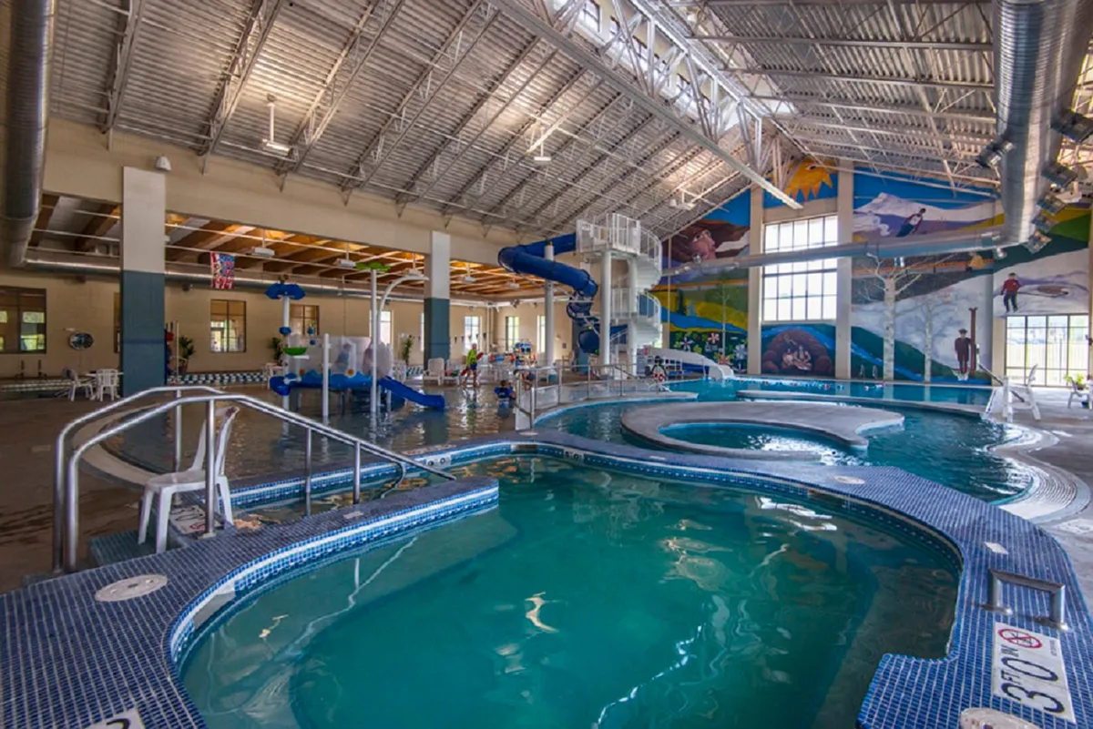 This interior shot showcases the lively pool area of the Grand Park Rec Center, featuring vibrant water slides, multiple swimming pools, and colorful murals. Families and children are visible enjoying the water and having fun.