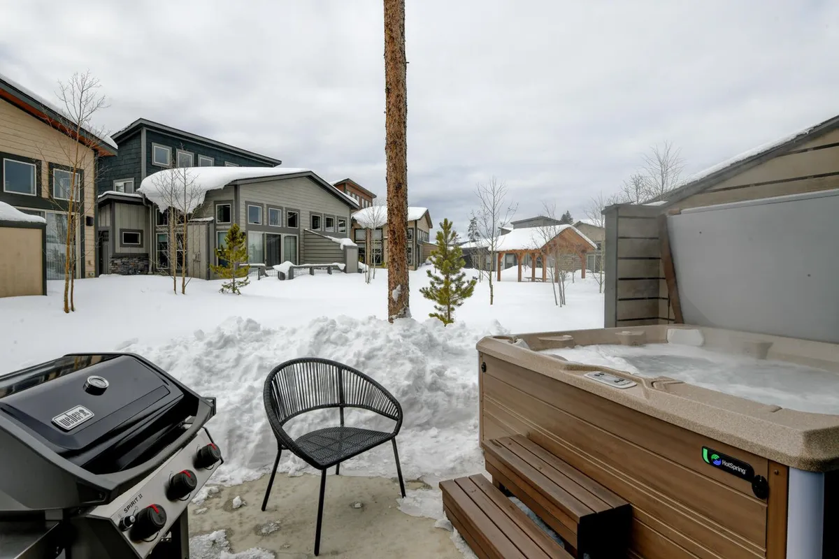 A cozy outdoor patio scene with a bubbling hot tub, grill, and comfortable seating, all set against a snowy landscape. The grill signals the potential for barbecues while the hot tub and chair emphasize relaxation, no matter the season.