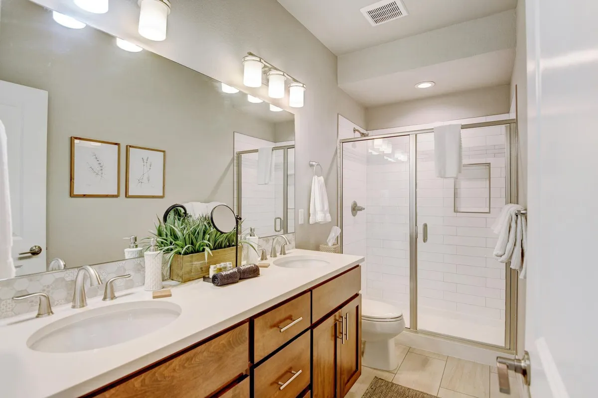 A modern bathroom featuring a double vanity with sinks, framed botanical prints on the walls, a glass shower enclosure, and a large vanity mirror. The room is bathed in natural light, emphasizing a spa-like, refreshing ambiance.