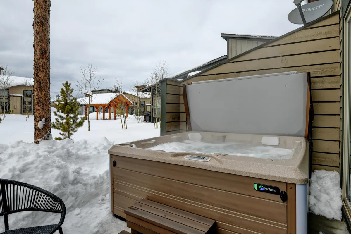 A serene winter scene featuring a private outdoor hot tub surrounded by snow-covered ground and tall trees. The hot tub is steaming, inviting relaxation, with space for multiple people to gather and enjoy the mountain air. The setting feels secluded yet welcoming, perfect for post-ski or hiking relaxation.